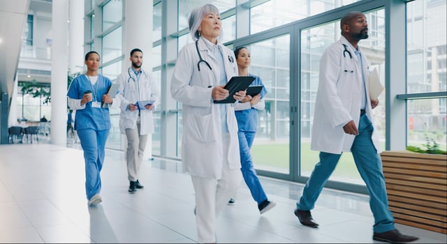 A group of doctors walking down a hallway, each wearing a white coat and carrying a clipboard.