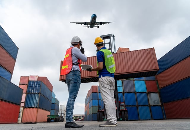 A man and a woman are standing in front of a large cargo ship, engaged in conversation. The woman is wearing a hard hat, and the man is holding a clipboard. The scene is set against a backdrop of a large airplane flying overhead.