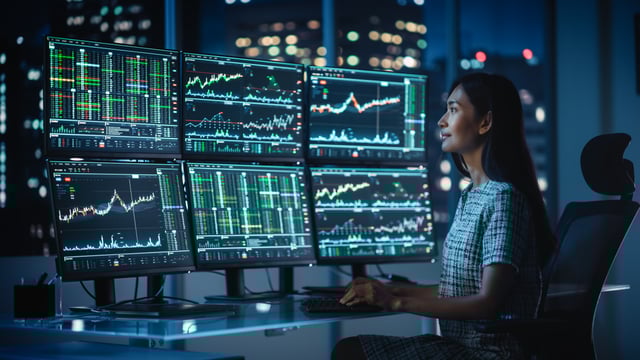 A woman is sitting at a desk in front of a computer screen displaying multiple graphs and charts. She appears to be working or analyzing the data displayed on the screens.